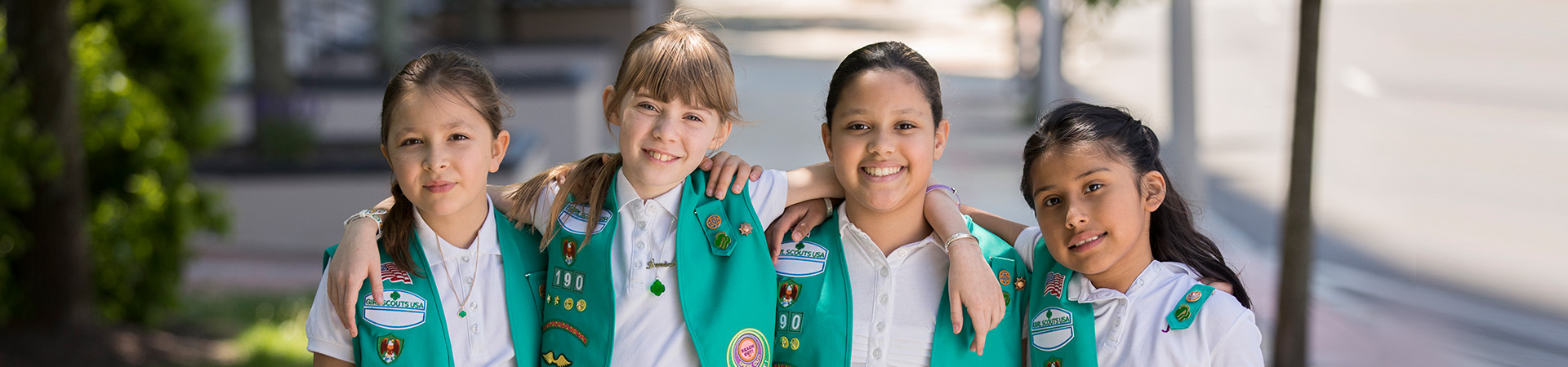  group of junior girl scouts walking outside hugging smiling at camera in junior vest and sash with badges 